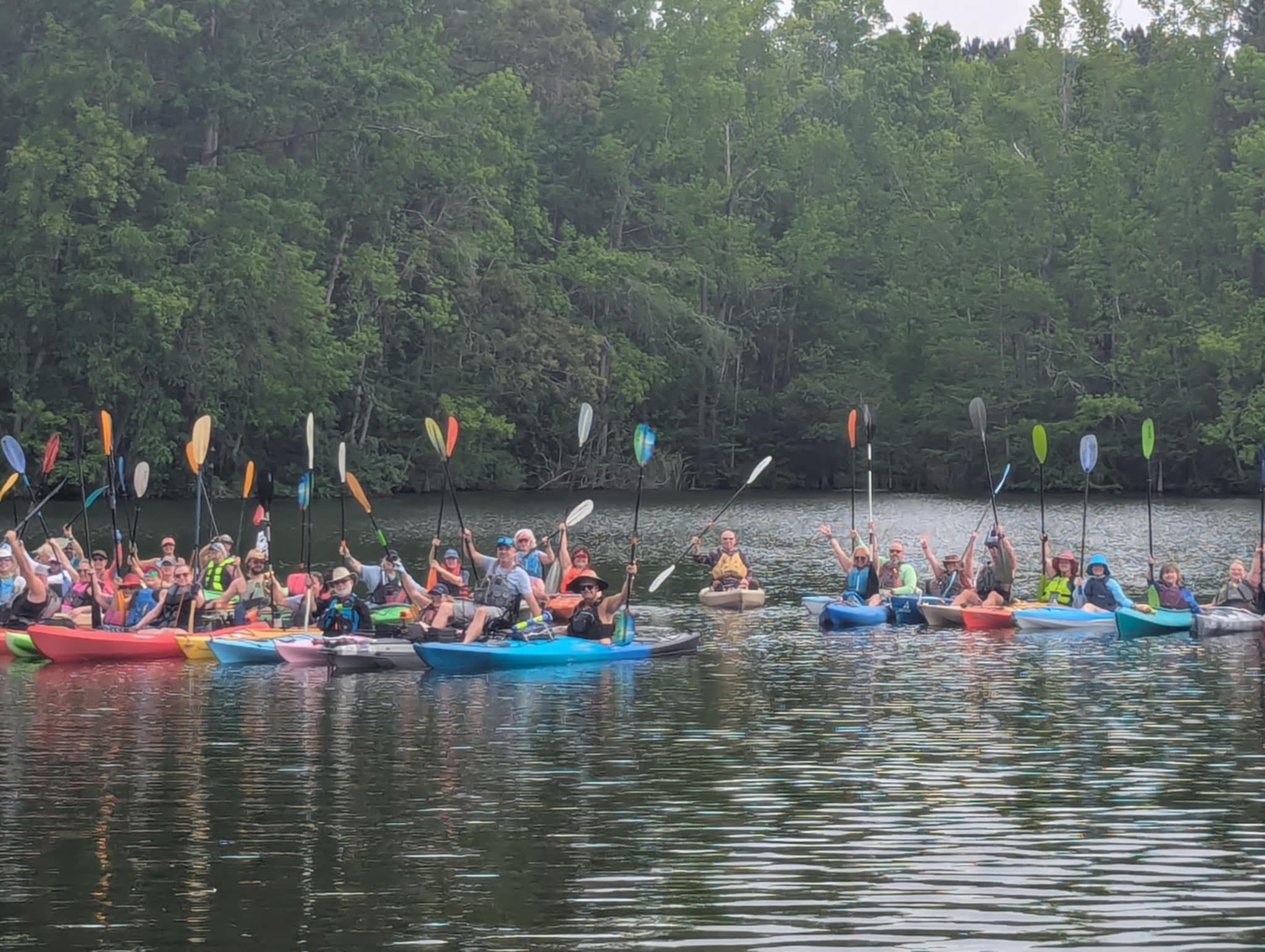 A large group of kayakers raising their paddles, from Dawn’s festival page